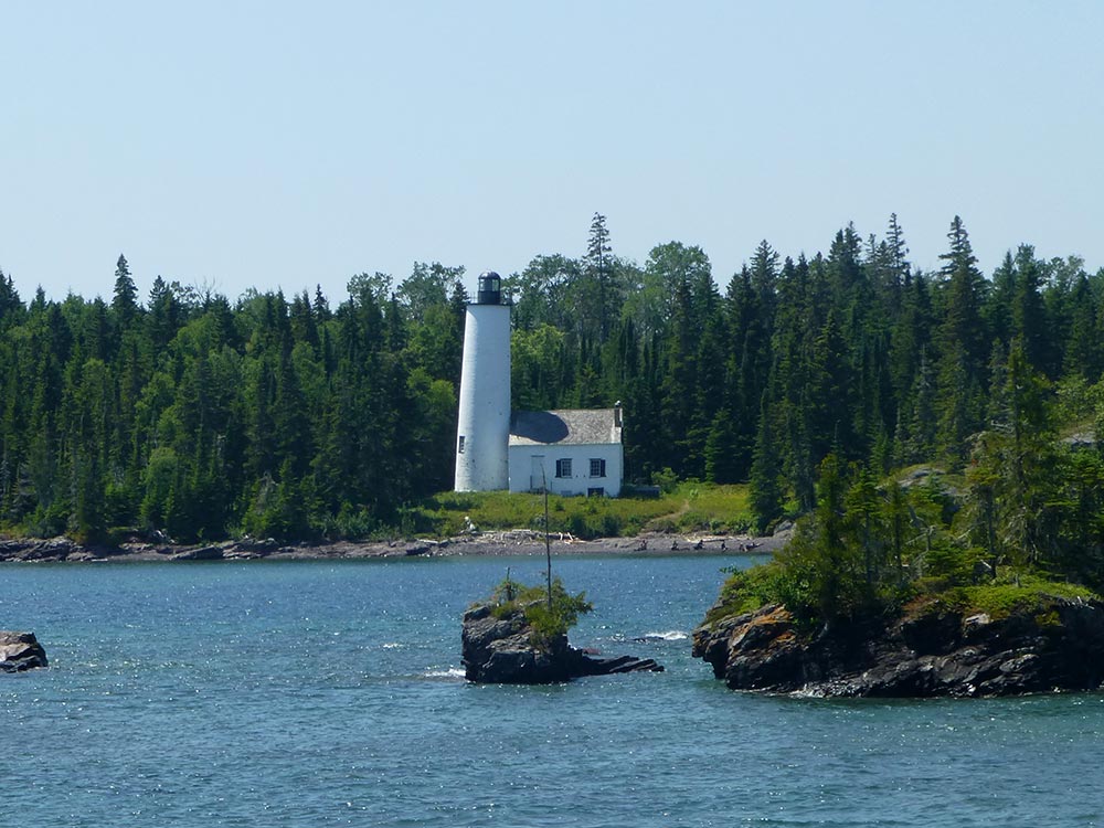 Rock Harbor lighthouse on Isle Royale. It is white, against the green backdrop of trees, with water in front. Seen from the Ranger III ferry.