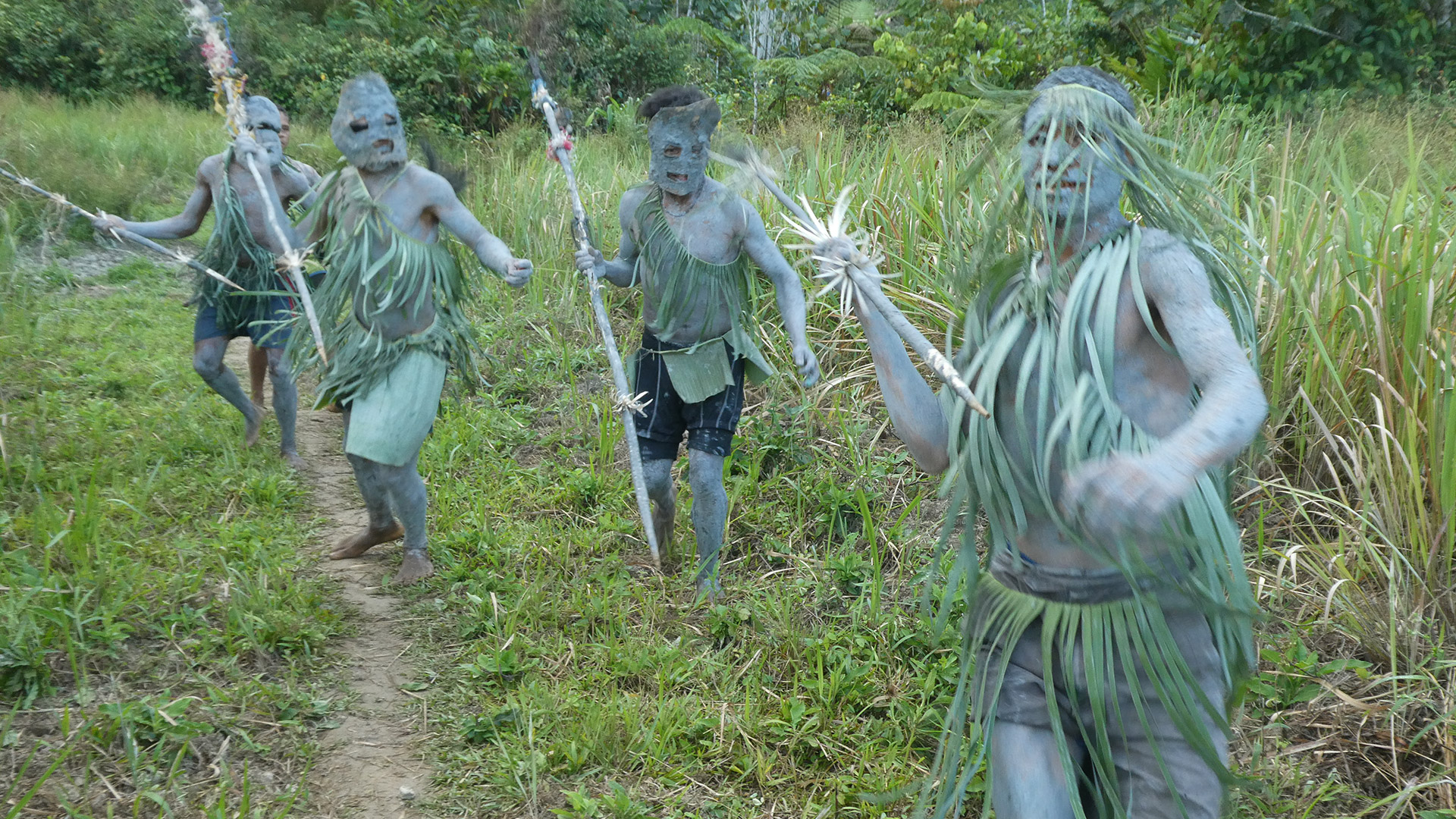 Men covered in mud and wearing leaves are running toward the camera with spears.