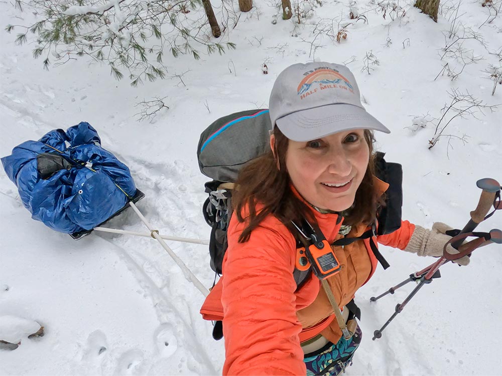 A woman looking up a a camera while smiling. She is hiking and pulling a pulk - a small sled with gear - behind her.