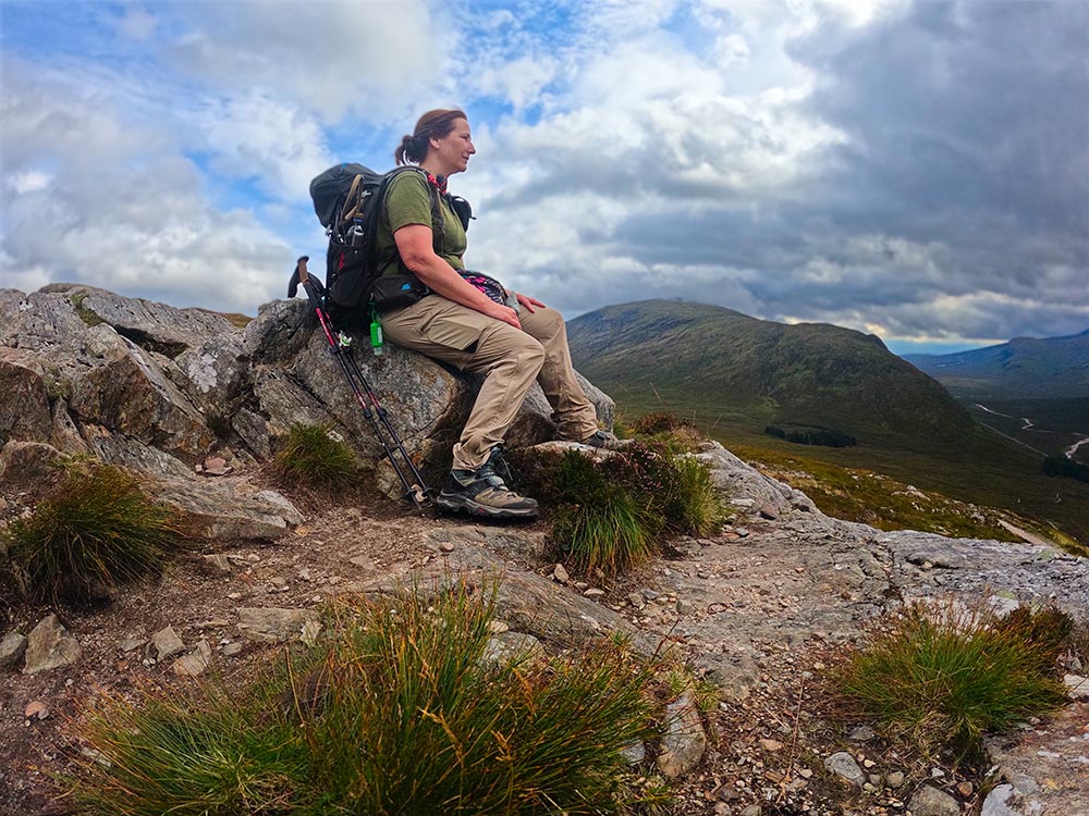 A woman sits on a rock atop a mountain looking into the distance - at the top of Devils Staircase, West Highland Way